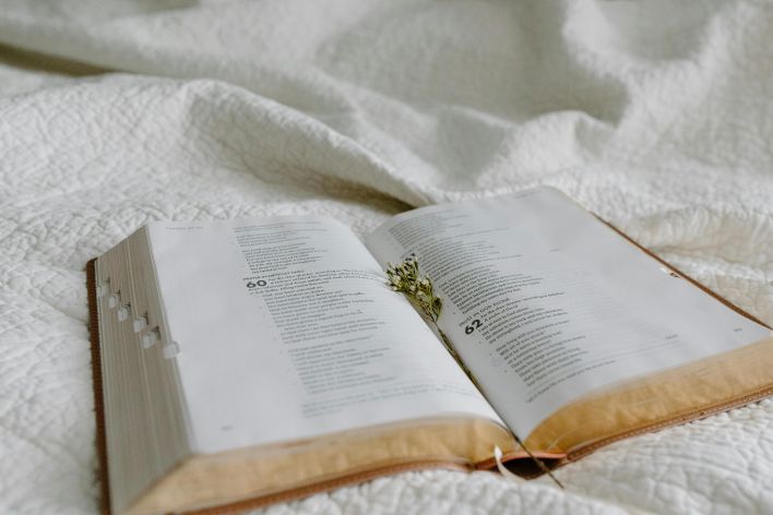An open Bible on a quilted bedspread with dried flowers marking a page, suggesting peaceful reflection.